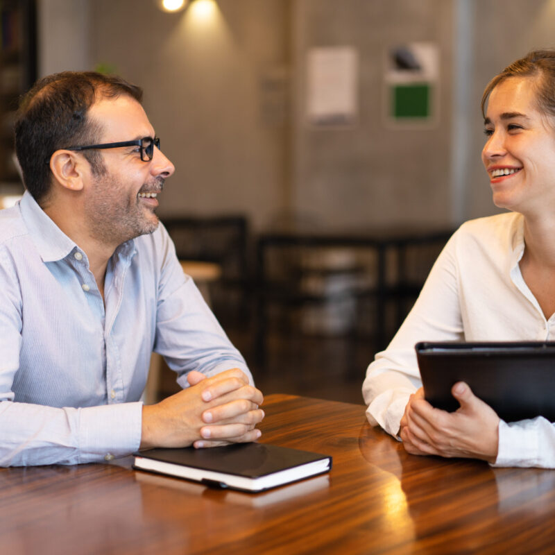 Happy mid adult manager talking to young female client. Caucasian executive wearing glasses sitting at table and talking to female colleague. Job interview or meeting with client concept