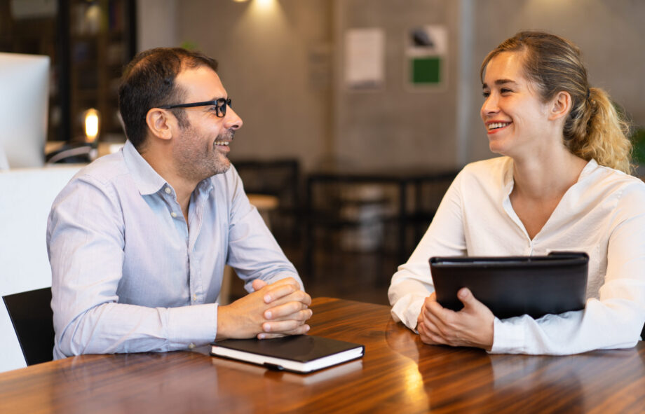 Happy mid adult manager talking to young female client. Caucasian executive wearing glasses sitting at table and talking to female colleague. Job interview or meeting with client concept